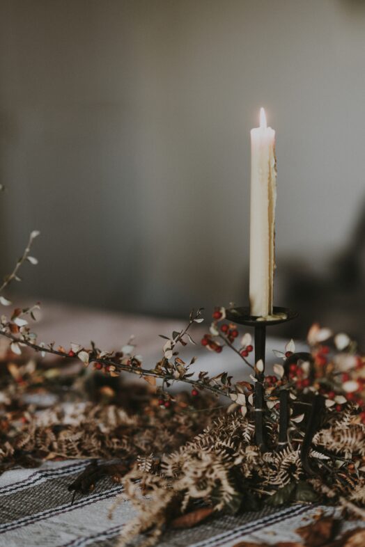 Ett ensamt tänt stearinljus står i en ljusstake, omgiven av torkade blommor på ett bord.